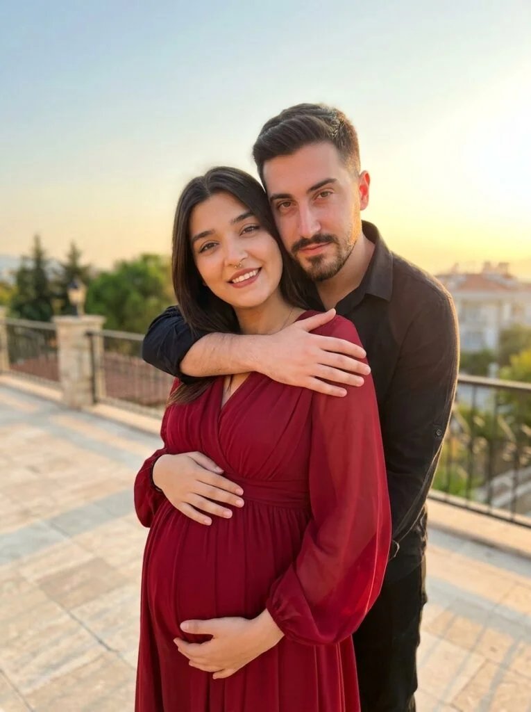 Expecting mother in a flowing red gown embraced by her partner at golden hour