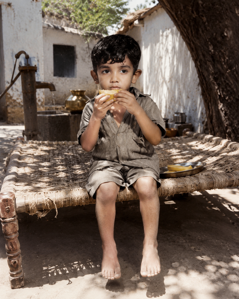 Child sitting on a charpai and eating mango at grandparents' house
